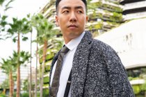 Joven emprendedor asiático bien vestido en corbata mirando hacia otro lado mientras pasea por la carretera contra edificios modernos en la ciudad - foto de stock