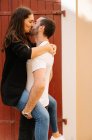 Vista lateral do romântico jovem étnico barbudo macho em roupas casuais carregando namorada feliz e beijando com os olhos fechados perto da porta na rua no dia ensolarado — Fotografia de Stock