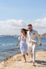 Ganzkörper fröhliches Barfuß-Hochzeitspaar läuft am Ufer in der Nähe des reißenden Meeres und genießt den Hochzeitstag in sonniger Natur — Stockfoto