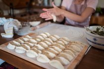 Женщина в фартуке катит тесто с руками на столе, готовя домашние пельмени на кухне — стоковое фото