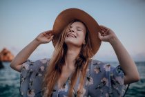 Счастливая женщина в стильном платье и шляпе стоит с закрытыми глазами и поднятыми руками на берегу моря в летний вечер — стоковое фото