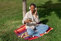 Full body of content African American female listening to music in wireless headphones while surfing cellphone on plaid near tree trunk in park — Stock Photo