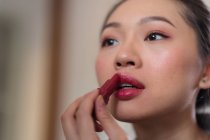 Cultiver jeune attrayant asiatique femelle en vêtements décontractés appliquant rouge à lèvres lumineux tout en regardant miroir — Photo de stock