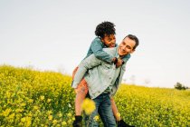 Romantic young man smiling and giving piggyback ride to joyful African American girlfriend in lush blooming yellow meadow in countryside — Stock Photo