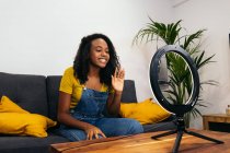 Smiling black female on couch waving hand while using smartphone on LED ring lamp near professional lights on tripods — Fotografia de Stock