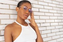 Selbstbewusste Afroamerikanerin mit kurzen Haaren in stylischem Outfit mit trendiger Brille steht auf der Straße in der Nähe der weißen Backsteinmauer — Stockfoto