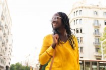 Femme afro-américaine positive dans des lunettes avec les cheveux noirs debout sur la rue avec des bâtiments résidentiels en ville contre ciel sans nuages — Photo de stock