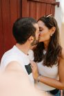 Vista laterale di innamorato giovane barbuto uomo etnico baciare fidanzata mentre prende selfie in piedi sulla strada nella giornata di sole — Foto stock