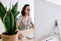 Щаслива жінка в повсякденному вбранні та окулярах, сидячи за столом з комп'ютером та переглядаючи смартфон — стокове фото