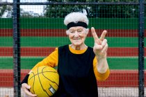 Оптимістична зріла жінка в активному одязі і пов'язка, дивлячись на камеру, стоячи на публічному баскетбольному майданчику з м'ячем під час тренувань — стокове фото