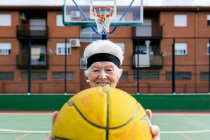 Mulher madura positiva em activewear e headband olhando para a câmera enquanto está de pé com bola durante o jogo de basquete — Fotografia de Stock