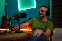 Positive junge bärtige männliche Millennial mit Brille und Kopfhörer lächelt und spricht in Mikrofon während der Aufnahme Podcast in dunklen Studio — Stockfoto