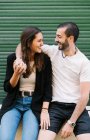 Alegre apaixonado jovem casal hispânico em roupas casuais rindo e olhando um para o outro, abraçando perto de parede verde na rua da cidade — Fotografia de Stock