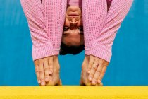 Mujer flexible en ropa deportiva de pie pose de flexión hacia adelante mientras practica la pose de yoga Uttanasana en la pared amarilla y azul - foto de stock
