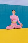 Mujer pacífica en ropa deportiva rosa sentada en Padmasana con las manos Namaste y meditando durante la sesión de yoga sobre fondo azul y amarillo - foto de stock