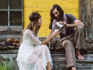 Hippie-Mann gießt Kaffee oder Tee aus einer Thermoskanne in eine Tasse, die von einer schönen Hippie-Frau auf einer Holzveranda gehalten wird — Stockfoto