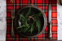 Dall'alto tavola natalizia allestimento con ghirlanda su piatto in ceramica con posate su tovaglia rossa a quadretti sullo sfondo — Foto stock