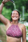 Felice femmina in piedi con gli occhi chiusi in cima e versando acqua dalla bottiglia sulla testa contro alberi verdi nel parco in estate — Foto stock