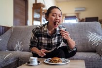 Молода азіатка їсть домашні млинці, які кладуть на тарілку біля чашки кави на стіл, сидячи на зручному дивані у вітальні. — стокове фото