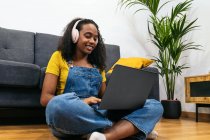 Femme noire souriante en salopette denim avec écouteurs assis sur le sol près du canapé et utilisant un ordinateur portable à la maison — Photo de stock