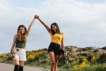 Glückliche multiethnische Freundinnen halten Händchen, während sie mit Longboards unterwegs sind — Stockfoto