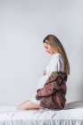 Seitenansicht einer jungen, sanften Schwangeren, die ihren Bauch berührt, während sie auf dem Bett sitzt — Stockfoto