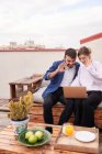 Casal positivo sentado perto e acenando as mãos para a câmera de netbook durante a conversa em vídeo no terraço perto da mesa com maçãs e suco — Fotografia de Stock