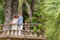 Junges Brautpaar in Hochzeitskostümen steht auf Holzsteg mit Geländer am See mit Felsen und grünen Palmen und Pflanzen im Park an Sommertagen — Stockfoto