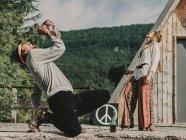 Full body side view of hippie couple singing and drinking alcohol drinks while resting together near wooden cabin in countryside — Stock Photo
