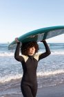 Side view of young happy female surfer in wetsuit with surfboard standing holding surfboard above head looking away on seashore washed by waving sea — Stock Photo