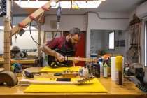 Mestre profissional em avental de pé perto da mesa com instrumentos e ferramentas enquanto ajusta guitarra elétrica na garagem moderna — Fotografia de Stock