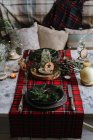 Christmas table setting with wreath on the plate, decorative wooden ornaments and red checkered tablecloth with yellow lights on the background — Stock Photo