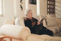 Focused senior female with gray hair resting on couch and reading e book on tablet in living room at home — Stock Photo