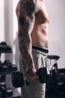 Vista lateral da cultura fisiculturista irreconhecível com tatuagens de pé com halteres pesados durante o treino no ginásio — Fotografia de Stock
