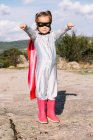 Corpo inteiro de menina pequena em traje de super-herói levantando punhos estendidos para mostrar poder enquanto estava em pé na colina rochosa — Fotografia de Stock