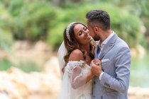 Seitenansicht des glücklichen jungen Ehepaares in Hochzeitskleidung stehend und Händchenhaltend in der Nähe des Sees und der grünen Bäume tagsüber im Garten — Stockfoto
