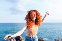 Feliz cabelo encaracolado fêmea estendendo os braços enquanto desfruta de liberdade na costa montanhosa da costa olhando para a câmera — Fotografia de Stock
