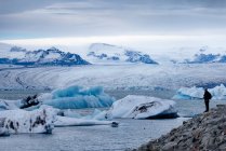Бічний вид далекого анонімного мандрівника в теплому верхньому одязі і капелюсі стоїть на скелястому пагорбі і захоплюється захоплюючим пейзажем Йокулсарлонського великого льодовикового озера під час поїздки в Ісландію. — стокове фото