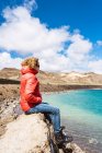 Вид сбоку спокойного человека, сидящего на краю скалы над голубым океаном на фоне гор в Исландии в солнечный день — стоковое фото