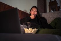Молода азіатка, одягнена в повсякденний одяг, сидить на дивані і їсть попкорн вдома, дивлячись кіно на ноутбуці. — стокове фото