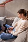Side view of young female sitting with crossed legs using modern netbook on comfortable couch on balcony in sunny day — Stock Photo