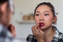Crop young attractive Asian female in casual clothes applying bright lipstick while looking at mirror — Stock Photo