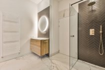 Просторная стеклянная душевая кабина и светящееся овальное зеркало, висящее на стене над раковиной, в просторной современной ванной комнате с мраморным полом — стоковое фото