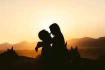 Seitenansicht von Silhouetten von liebenden Pärchen, die sich umarmen, während sie Zeit miteinander auf der Weide in der Nähe von Pferden vor Bergen im Sonnenuntergang verbringen — Stockfoto
