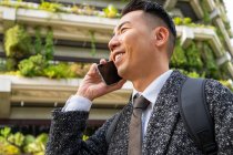 Alegre joven empresario étnico masculino con corbata mirando hacia adelante mientras habla por teléfono celular en la ciudad - foto de stock