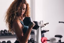 Вид сбоку сильной женщины с кудрявыми волосами, делающей упражнения на бицепсах с гантелями во время тренировки в спортзале — стоковое фото
