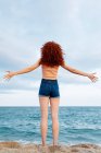 Back view full length of unrecognizable barefoot female traveler standing on sandy coast washed by foamy waves of blue sea — Stock Photo