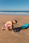 Full body side view of active male in swimming shorts attaching leg corde while preparing for surfing on sandy beach near sea — Photo de stock