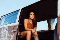 Atractiva chica morena con sombrero dentro de una furgoneta vintage y tumbada en el asiento en un día soleado - foto de stock