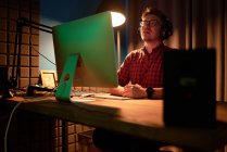 Концентрований самець у картатій сорочці та окулярах працює на комп'ютері, сидячи за столом з лампою та мікрофоном під час запису подкасту — стокове фото